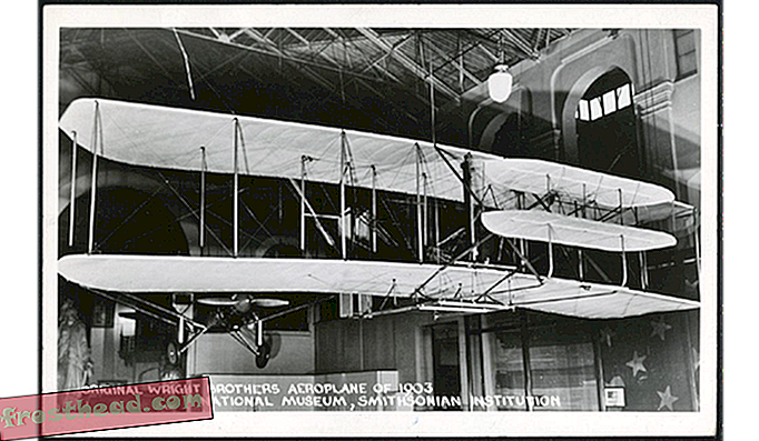 Černobílá fotografická pohlednice zobrazuje letoun Wright Flyer z roku 1903, který visí na displeji v budově Arts and Industries Building, kde byl na pohled od roku 1948 do roku 1976, než byl přesunut do Národního muzea pro vzduch a vesmír. Foto s laskavým svolením Smithsonian Ins